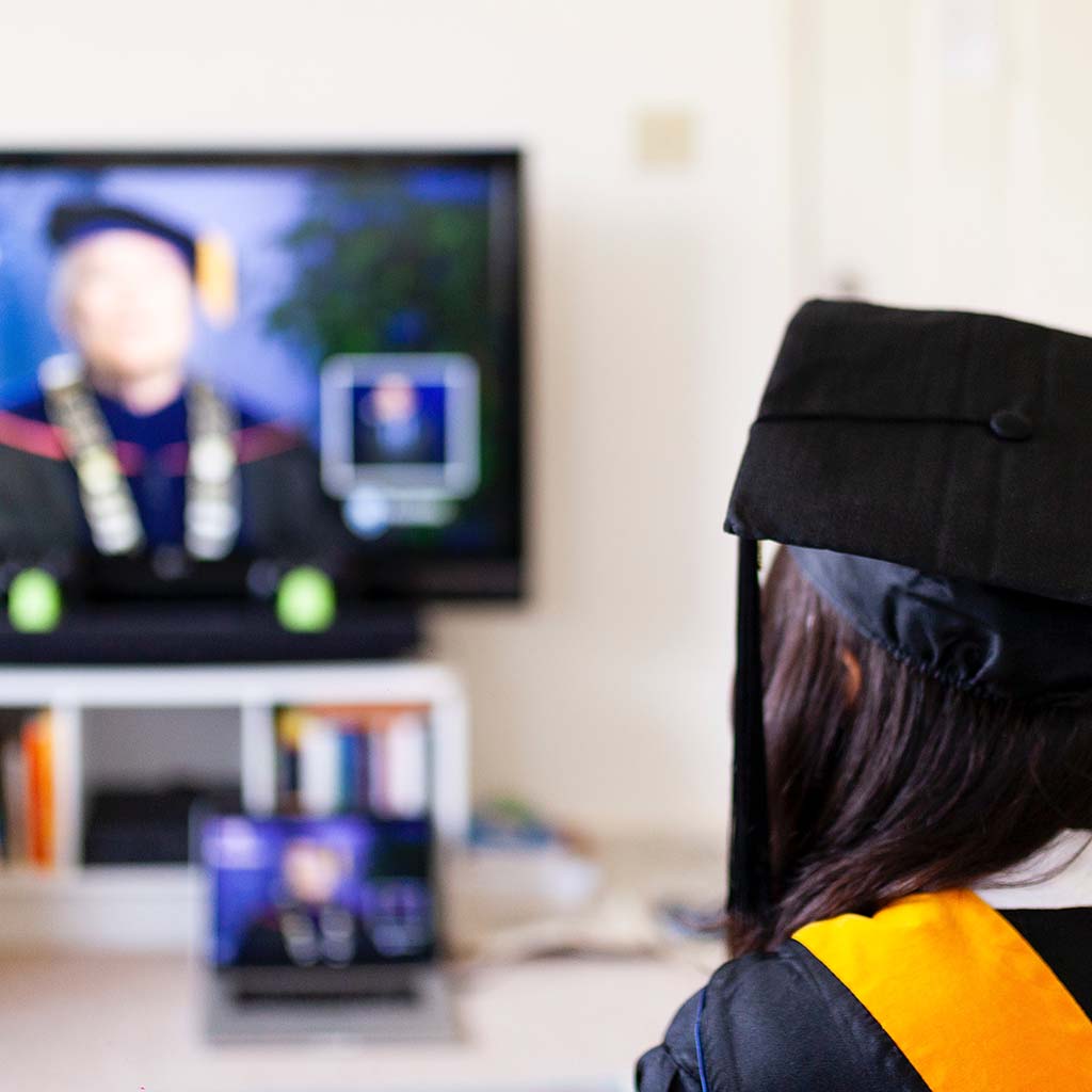 Student in cap and gown watching teleconference on laptop.
